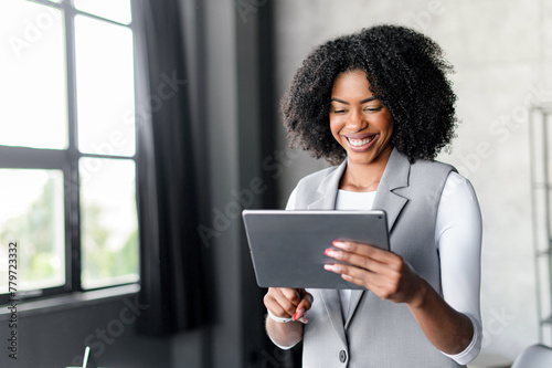 An African-American businesswoman in a grey suit smiles while engaging with her tablet, standing in an office. This shot emphasizes dynamic business activity and the seamless integration of technology