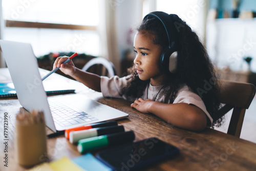 Adorable African American little girl sitting at wooden table on laptop computer and listening online school class over headphones while looking at screen and doing home work in living room photo