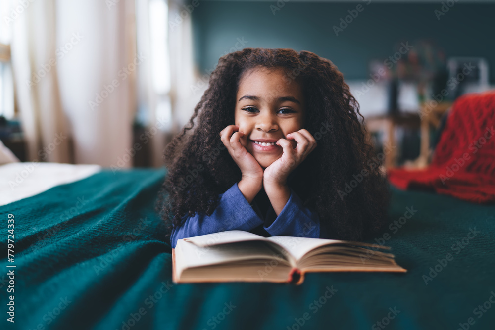 Joyful African American girl, aged 6-8, lying on her stomach on a green blanket, hands on cheeks, with a book open in front of her. Her bright smile and sparkling eyes, paired with a blue top - obrazy, fototapety, plakaty 