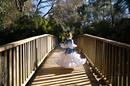 A pretty girl dancing flamenco in a typical gypsy dress with frills and fringes walks on a wooden bridge in a famous park in Seville, Spain. Her hair is tied back with a flower.
