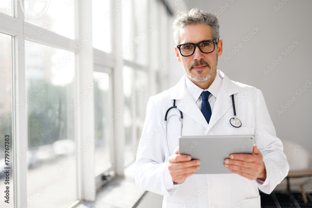 A professional senior doctor with grey hair is standing in a well-lit office, holding a digital tablet in hands, looking at the camera with serious face expression A professional senior doctor with grey hair is standing in a well-lit office, holding a digital tablet in hands, looking at the camera with serious face expression