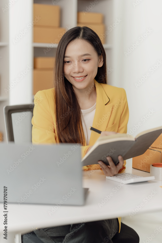 Freelance Asian woman wearing an apron uses laptop and boxes to take and check online orders to prepare packs to sell to customers. SME online business idea, vertical image Freelance Asian woman wearing an apron uses laptop and boxes to take and check online orders to prepare packs to sell to customers. SME online business idea, vertical image