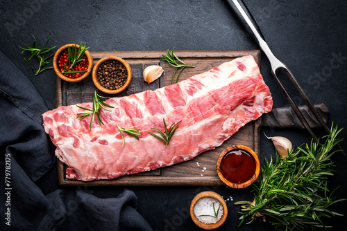 Raw pork ribs with rosemary, spices and barbecue sauce on oak wooden cutting board prepared for cooking on black kitchen table background, top view