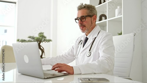 A genial mature doctor in a white coat waves hello into laptop during online appointment, seated in a well-organized clinic office that reflects a clean and efficient medical environment, taking notes