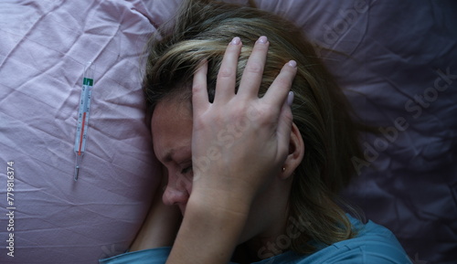 Young woman in bed and holding her head near mercury thermometer. Symptoms of bacterial meningitis concept photo