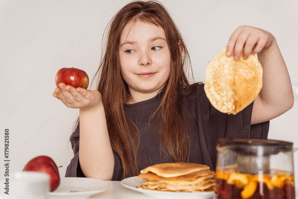 A little teenage girl holds a pancake in one hand and a red apple in the other and looks at it. Concept of choosing the right food. High quality photo A little teenage girl holds a pancake in one hand and a red apple in the other and looks at it. Concept of choosing the right food. High quality photo