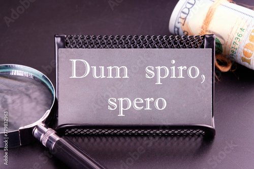 Dum Spiro Spero - latin phrase means While I Breath, I Hope. on the business card next to a roll of money and a magnifying glass on a black background photo