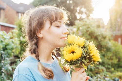 Dreaming young female farmer woman with closed eyes holding and sniffing a sunflowers bouquet on the green garden background in sunset light. Enjoy the moment. Rural, Cottage core lifestyle. photo