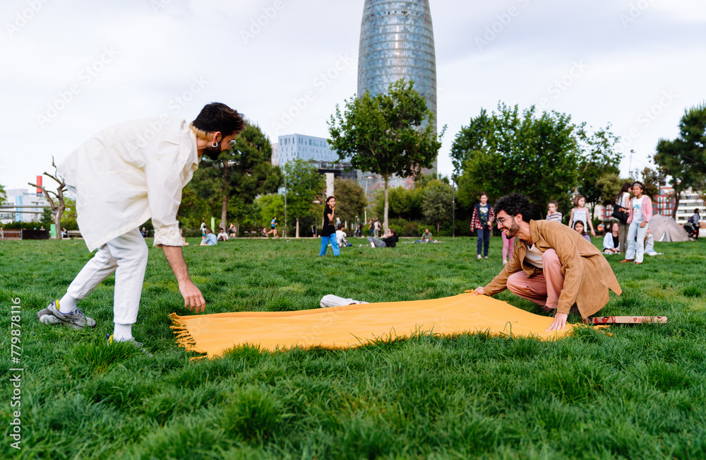 Young male couple spreading blanket on meadow in park