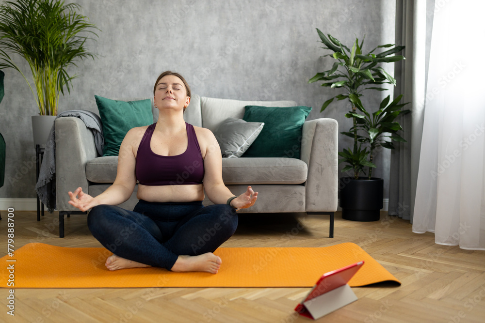 Sporty plus size woman in sportswear working out at home. Sports, activity and weight loss concept. Young plus size woman in leggings exercising on the mat, watching online video via tablet Sporty plus size woman in sportswear working out at home. Sports, activity and weight loss concept. Young plus size woman in leggings exercising on the mat, watching online video via tablet