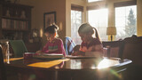 Two siblings at the dining room table, working on homework together. The late afternoon sunlight streams in, casting long, soft shadows and a warm glow over their shared learning e