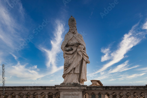 Stone statue of religious figure in Vatican City, adorned in ecclesiastical garments, holding a book. Against blue sky with clouds, hints at grandeur.