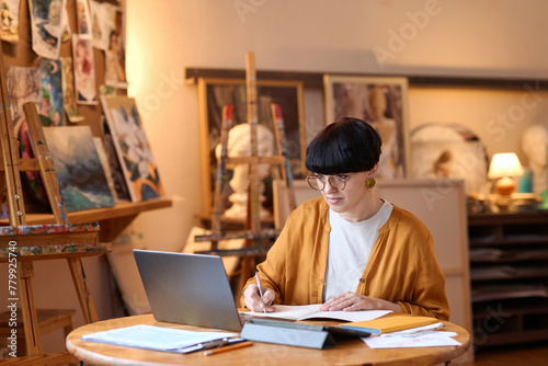 Wide angle view of funky female artist using laptop in art studio and taking notes while managing small business