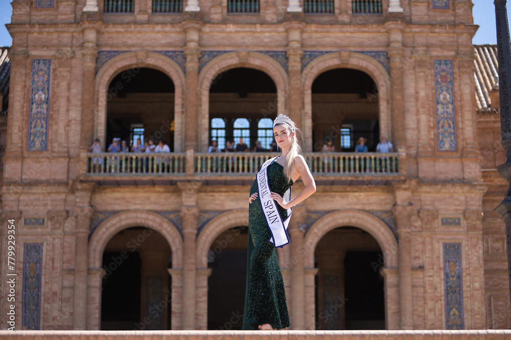 Young, pretty, blonde woman in a green party dress with sequins, with a diamond crown and beauty pageant winner's sash, posing in the middle of a square. Concept of beauty, fashion, trend, elegance.