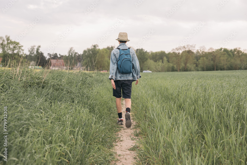 Back view of a teenage girl with straw hat, denim jacket and backpack on her back walking along a trail in the field