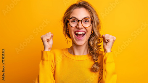 A woman in a yellow sweater is smiling and raising her hands in the air. She is wearing glasses Photo of ecstatic lady shout loud yeah fist up raise win lottery isolated bright shine color background photo