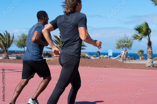 Two healthy sportsmen running outdoors along beach with tropical palm trees on hot summer day with clear blue sky, male friends jogging in morning during vacation. Healthy life and wellness concept