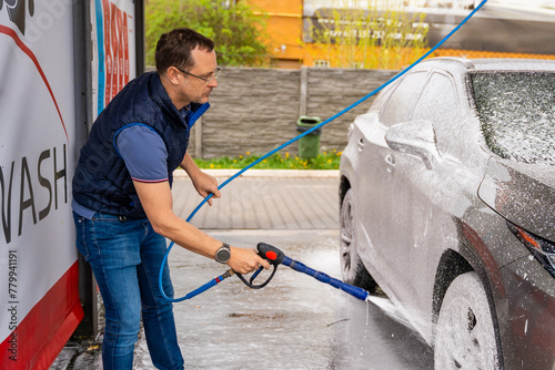 Young man washes his car at a self-service car wash using a hose with pressurized water and foam. photo