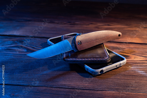 A sharp folding knife lying on a wallet and smartphone. A sharp knife with a wooden handle.