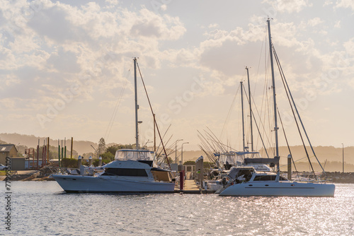 Tall masts on a row of yachts moored at a coastal marine with hazy clouds over sparkling water