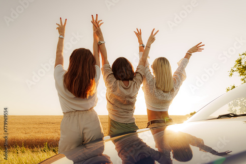 Three joyful women raise their hands in celebration of freedom during a road trip at sunset, with a vast field in the background.