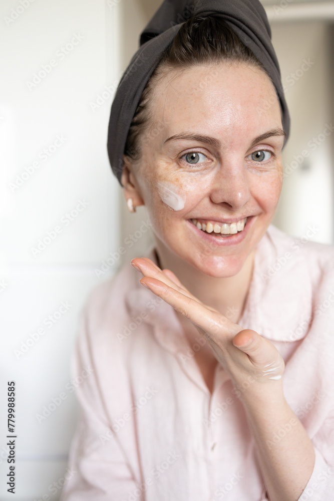 Woman in turban after washing hair looking at camera, smiling and showing cosmetic cream smear on face. Concept of morning skincare routine, anti-age care, hydration, no retouch Woman in turban after washing hair looking at camera, smiling and showing cosmetic cream smear on face. Concept of morning skincare routine, anti-age care, hydration, no retouch