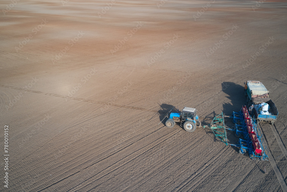  The process of replenishing the seeder compartments with seeds and mineral fertilizer. Panoramic aerial view of a tractor with a seeder and a truck with a load. Two workers are loading the seeder. 