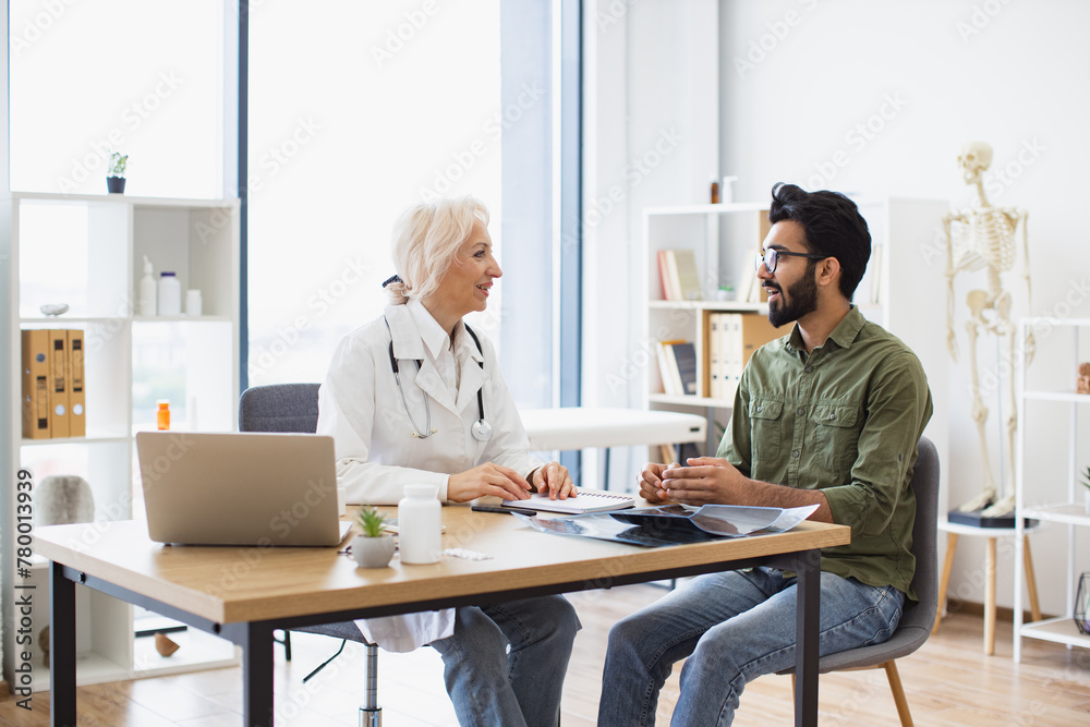 Bearded gentleman describing symptoms of illness to senior female doctor and gesturing hands. Mature elder physician giving recommendations and using notebook for writing prescription.