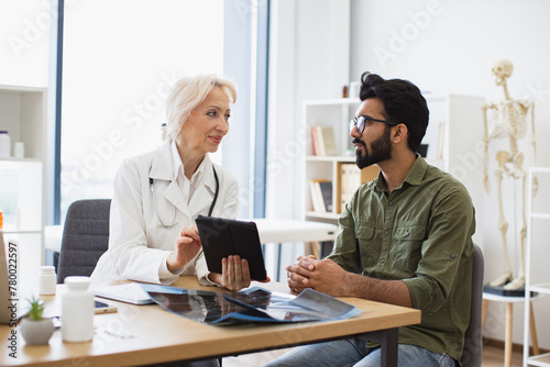 Young gentleman describing symptoms of illness to female doctor. Gray haired physician giving recommendations and using tablet for writing electronic prescription. © sofiko14
