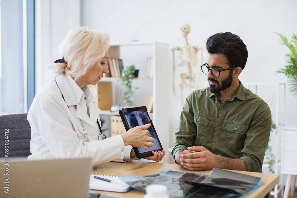 Family doctor examining diagnostic test results while developing treatment plan. Mindful gray-haired female in lab coat holding tablet with MRI scan while brunette guy sitting next to her at desk.