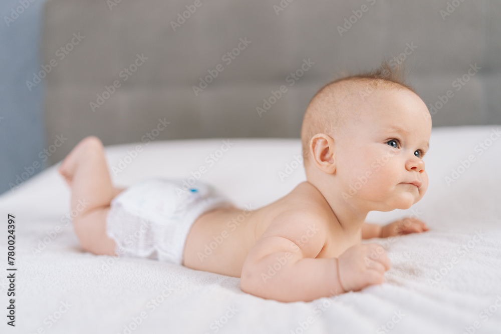 Medium shot of sweet infant baby wearing white diaper lying on stomach and happily waves arms and legs with smile. Portrait of nice newborn child learning to hold head on comfortable blanket at home. Medium shot of sweet infant baby wearing white diaper lying on stomach and happily waves arms and legs with smile. Portrait of nice newborn child learning to hold head on comfortable blanket at home.