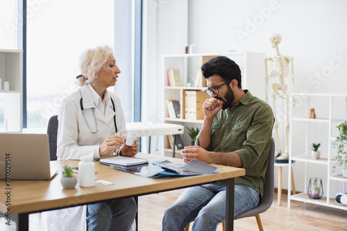 Elder female doctor providing expert check-up and giving medicine to young patient in hospital office. Focused man listening attentively to recommendations regarding medical history kept on laptop.