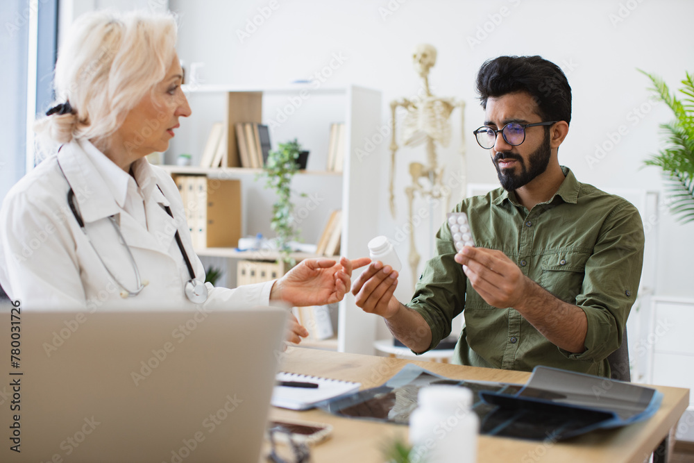 Doctor discussing health issues and reviewing past medical data on laptop. Senior female general practitioner communicating with young patient holding bottle and blister of medication in new clinic. Doctor discussing health issues and reviewing past medical data on laptop. Senior female general practitioner communicating with young patient holding bottle and blister of medication in new clinic.