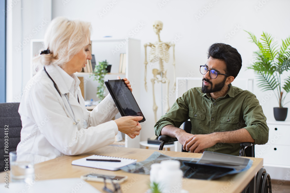 Attractive bearded young man looking on tablet with x-ray image carried by radiologist in consulting room. Confident senior specialist discussing results of diagnostic tests with wheelchair user. Attractive bearded young man looking on tablet with x-ray image carried by radiologist in consulting room. Confident senior specialist discussing results of diagnostic tests with wheelchair user.