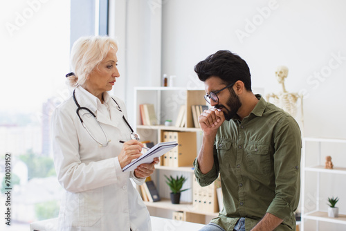 Elder specialist writing instruction for taking medication correctly. Caucasian senior female doctor and young male patient sitting at exam couch of modern medical center and diagnose disease.