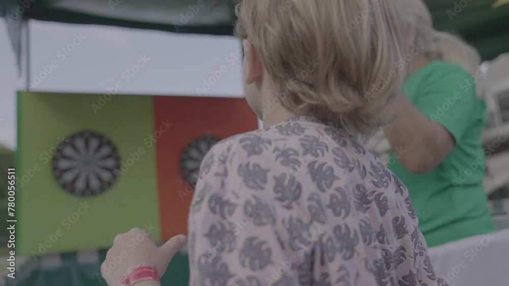 Niño rubio juega al tiro al blanco tirando un dardo en una feria o kermesse