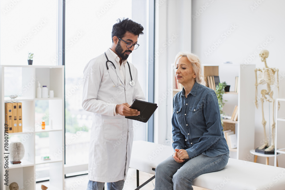 Bearded male doctor and elderly female patient sitting at exam couch of modern medical center and diagnose disease. Young specialist writing instruction for taking medication correctly.