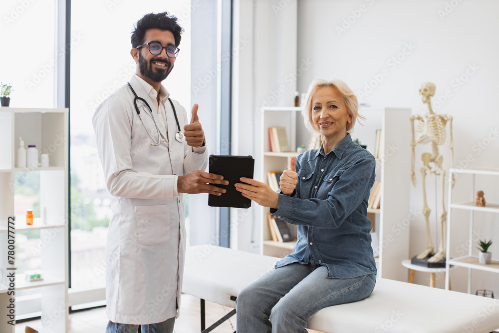Fototapeta premium Portrait of bearded physician using tablet for writing electronic prescription. Senior woman describing symptoms of illness to young male doctor at modern medical office, looking, at camera.