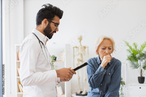 Bearded specialist writing electronic instruction for taking medication correctly. Respected man doctor and senior woman patient sitting at exam couch of new medical center and diagnose disease.
