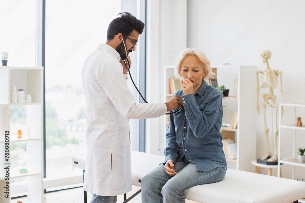 Bearded man in white lab coat checking lungs and heart via stethoscope. Serious old woman in everyday wear, coughing while visiting family doctor for full examination in general practice. Bearded man in white lab coat checking lungs and heart via stethoscope. Serious old woman in everyday wear, coughing while visiting family doctor for full examination in general practice.