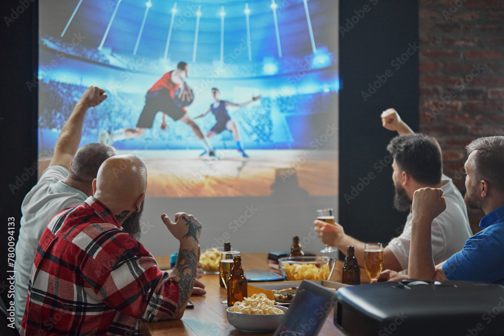 Men, friends sitting at table with beer and snacks, watching online basketball game translation on TV and cheering up favorite team. Concept of sport, championship, game, sport fans, leisure - obrazy, fototapety, plakaty 