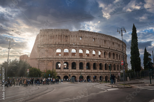 Iconic ancient amphitheater in Rome, Italy shows Colosseum's grandeur with multiple arches and historical architecture. Overcast sky, modern elements, tourists, pedestrians nearby.