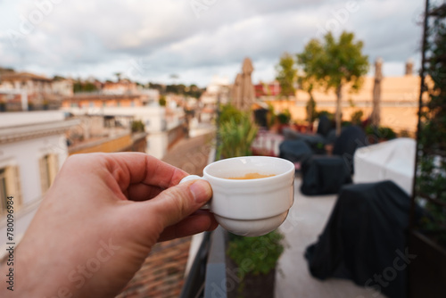 Photography Luxury terrace in Rome close up of hand holding white espresso cup, rooftops of warm colored buildings in the background, hints of greenery and overcast sky