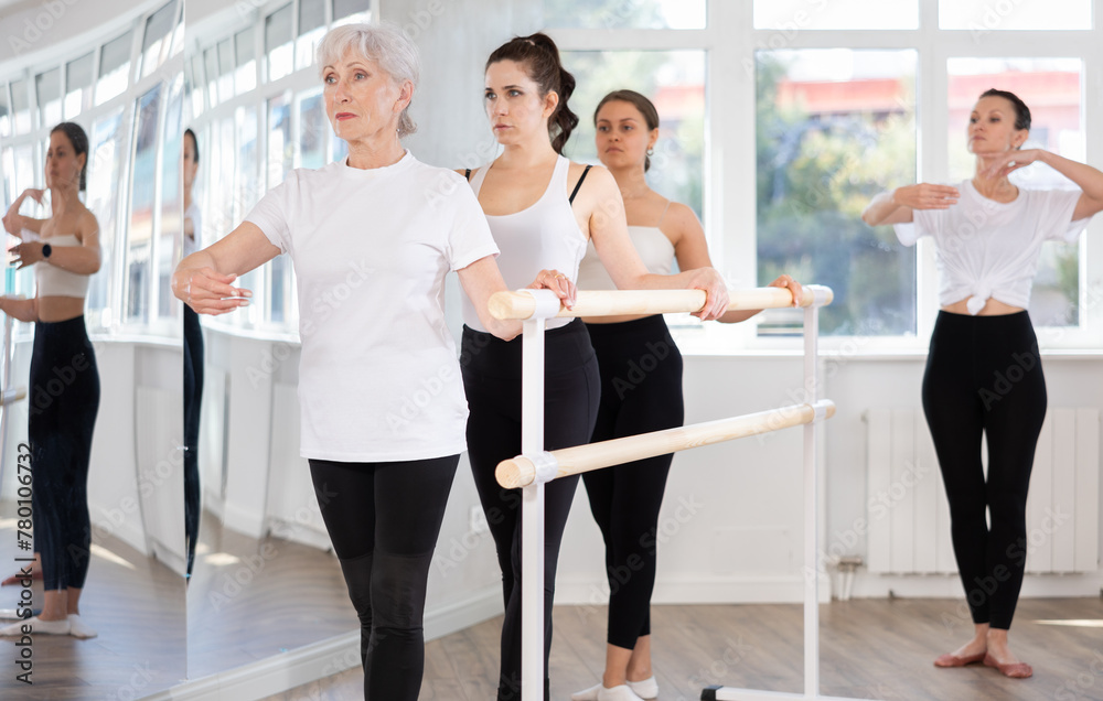 Concentrated graceful elderly woman practicing basic ballet moves at barre during multi-age group class under guidance of experienced female instructor