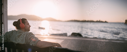Portrait of a young man in bright large headphones listening to music, audiobook by the sea at sunset. photo