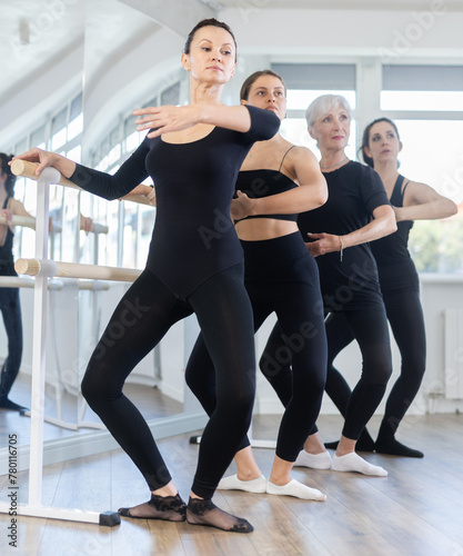 Concentrated woman practicing demi plie at barre during group ballet class for beginners under guidance of experienced elderly female instructor..