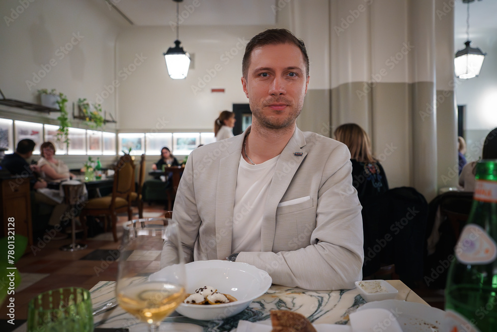 Man in light blazer at elegant restaurant table in Rome hotel, smiling at camera. Plate with remains of a meal, wine and water. Classic decor, warm ambiance, lively atmosphere. Man in light blazer at elegant restaurant table in Rome hotel, smiling at camera. Plate with remains of a meal, wine and water. Classic decor, warm ambiance, lively atmosphere.