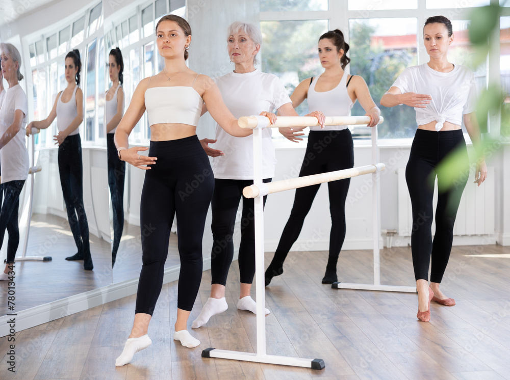 Fototapeta premium Concentrated graceful young girl practicing battement tendu at barre during ballet class for beginners with group of women of different ages..