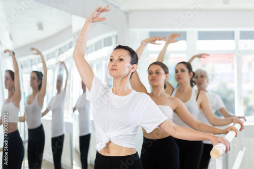 Enthusiastic fit woman maintaining third position at ballet barre at group rehearsal, mastering balletic technique and posture in mirrored choreography studio