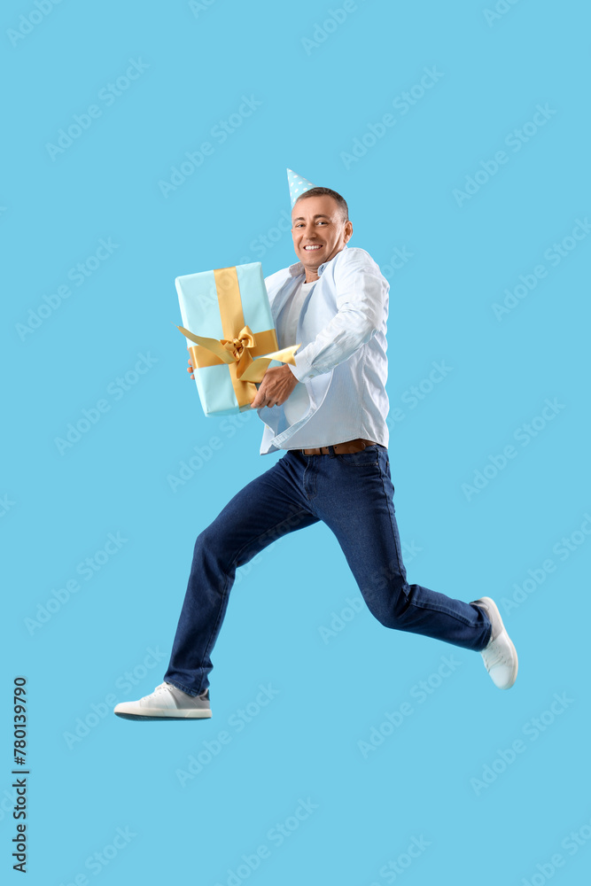 Mature man in party hat with birthday gift jumping on blue background Mature man in party hat with birthday gift jumping on blue background